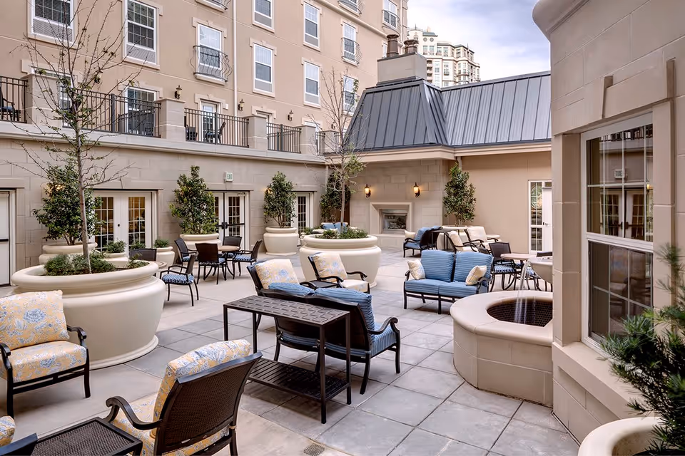 Outdoor courtyard area at Belmont Village Senior Living Turtle Creek featuring multiple seating arrangements with cushioned chairs and sofas, large potted plants, a built-in fireplace, and a fire pit surrounded by beige walls and windows of the building.
