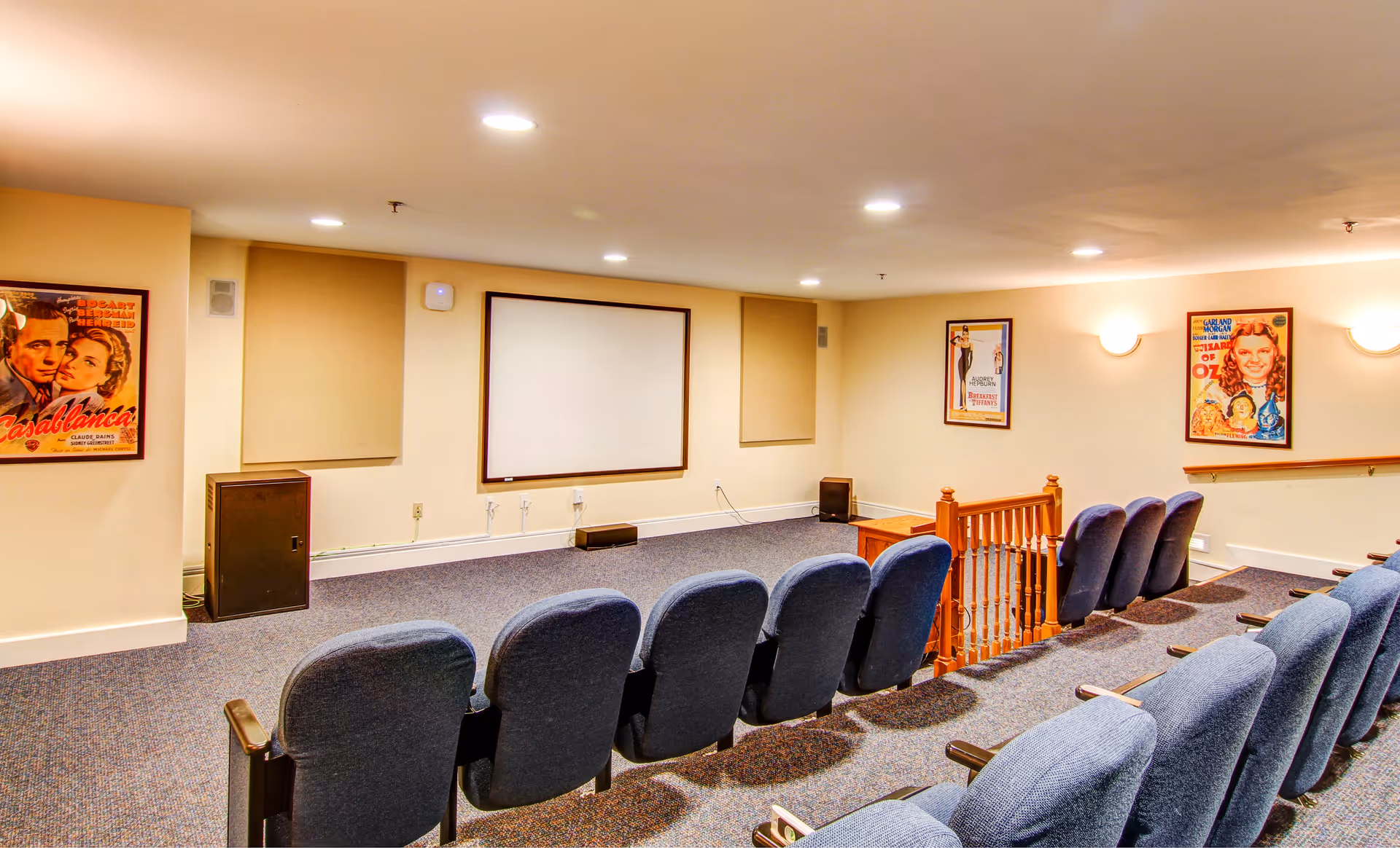 A small theater room with two rows of blue cushioned seats facing a blank white projection screen on a beige wall. The room has carpeted floors, framed vintage movie posters on the walls, and recessed ceiling lights. There is a wooden railing near the back row of seats.