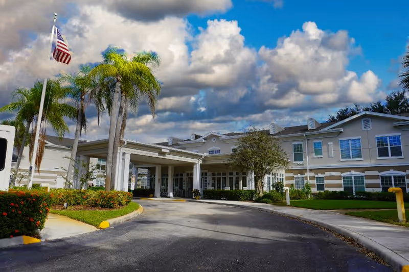 Senior living facility entrance with palm trees and an American flag under a cloudy sky.