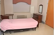 A simple bedroom with a single bed covered in a pink sheet, a wooden dresser with a mirror, a small table, a window with a closed blind, and a wooden door.