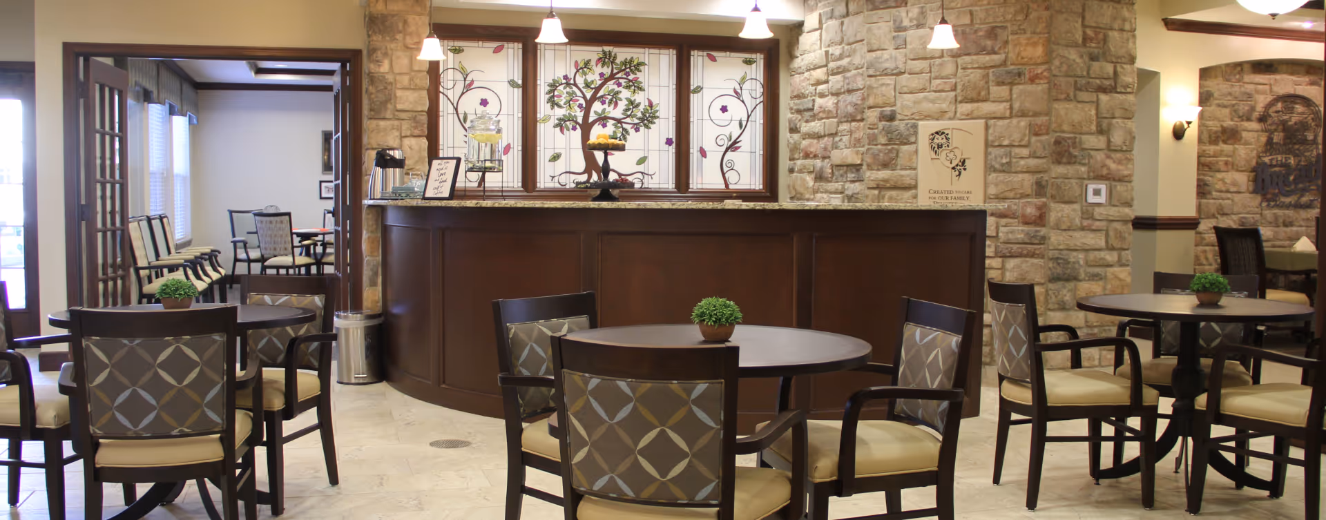 A cozy dining area in a senior living facility with round tables and cushioned chairs. The room features a stone wall with a counter topped with a glass container of lemon water and a tiered tray of fruit. Decorative stained glass windows with tree and floral designs are behind the counter. Pendant lights hang from the ceiling, and there are small potted plants on the tables.