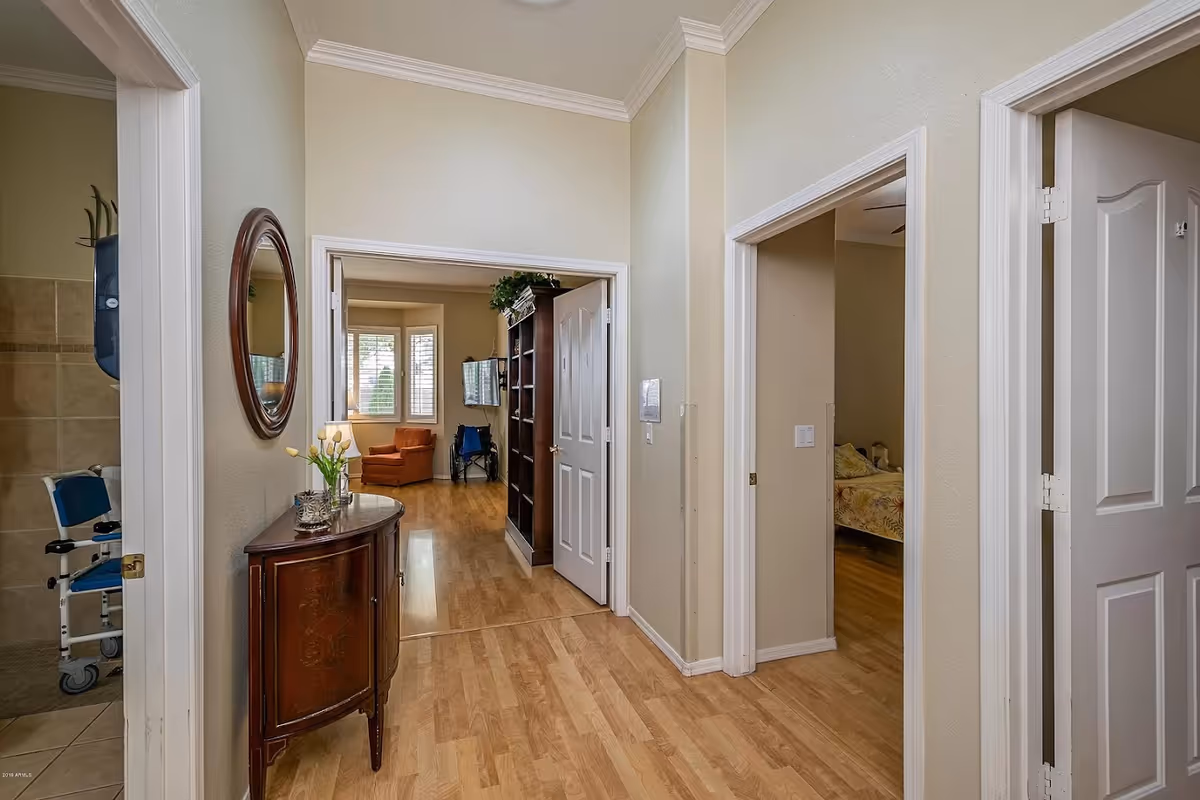 Interior hallway of a senior living facility with light wood flooring and beige walls. On the left, there is a bathroom with a shower chair visible. A wooden console table with a vase of yellow tulips and a round mirror hangs above it. Straight ahead is a living room area with an orange armchair, a wheelchair, a wall-mounted TV, and a large window with white shutters. To the right, there is a bedroom with a bed covered in floral bedding.