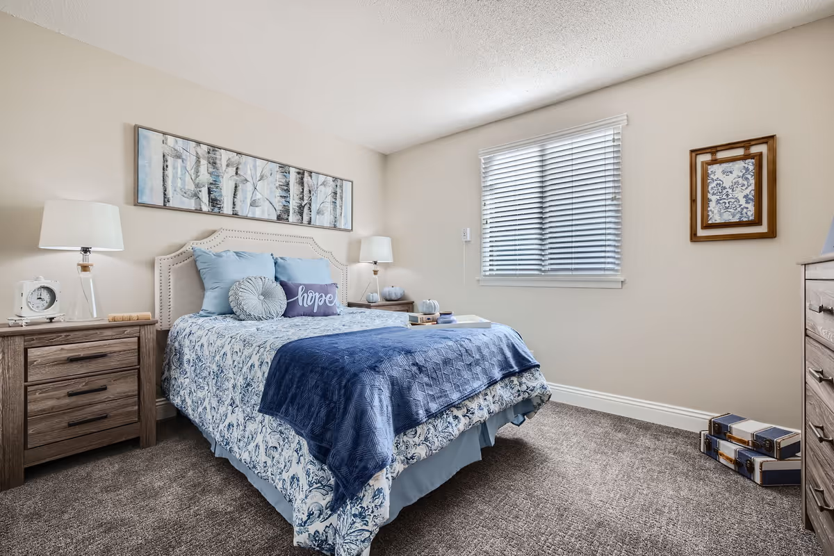 Bright, tastefully decorated bedroom featuring a bed with blue bedding, nightstands and lamps, a window with blinds, and framed artwork.