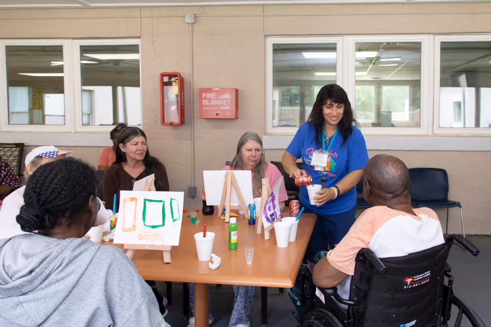 A group of elderly individuals seated around a table engaged in a painting activity. A caregiver in a blue shirt is pouring a drink into a cup for a man in a wheelchair. The table has small easels with canvases showing simple paintings, paint bottles, and cups. The setting appears to be an indoor common area with windows and a fire blanket mounted on the wall.