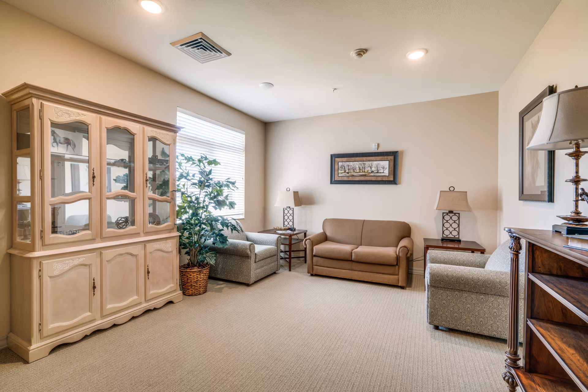 Neutral, well-lit sitting room with a sofa, armchairs, side tables with lamps, a glass-front cabinet, and a potted plant.