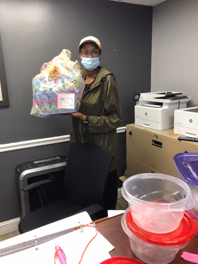 A person wearing a face mask, glasses, a cap, and a green jacket stands in an office holding a colorful gift basket wrapped in clear plastic. The office has gray walls, a black chair, a paper shredder, filing cabinets with printers on top, and plastic containers on a desk in the foreground.