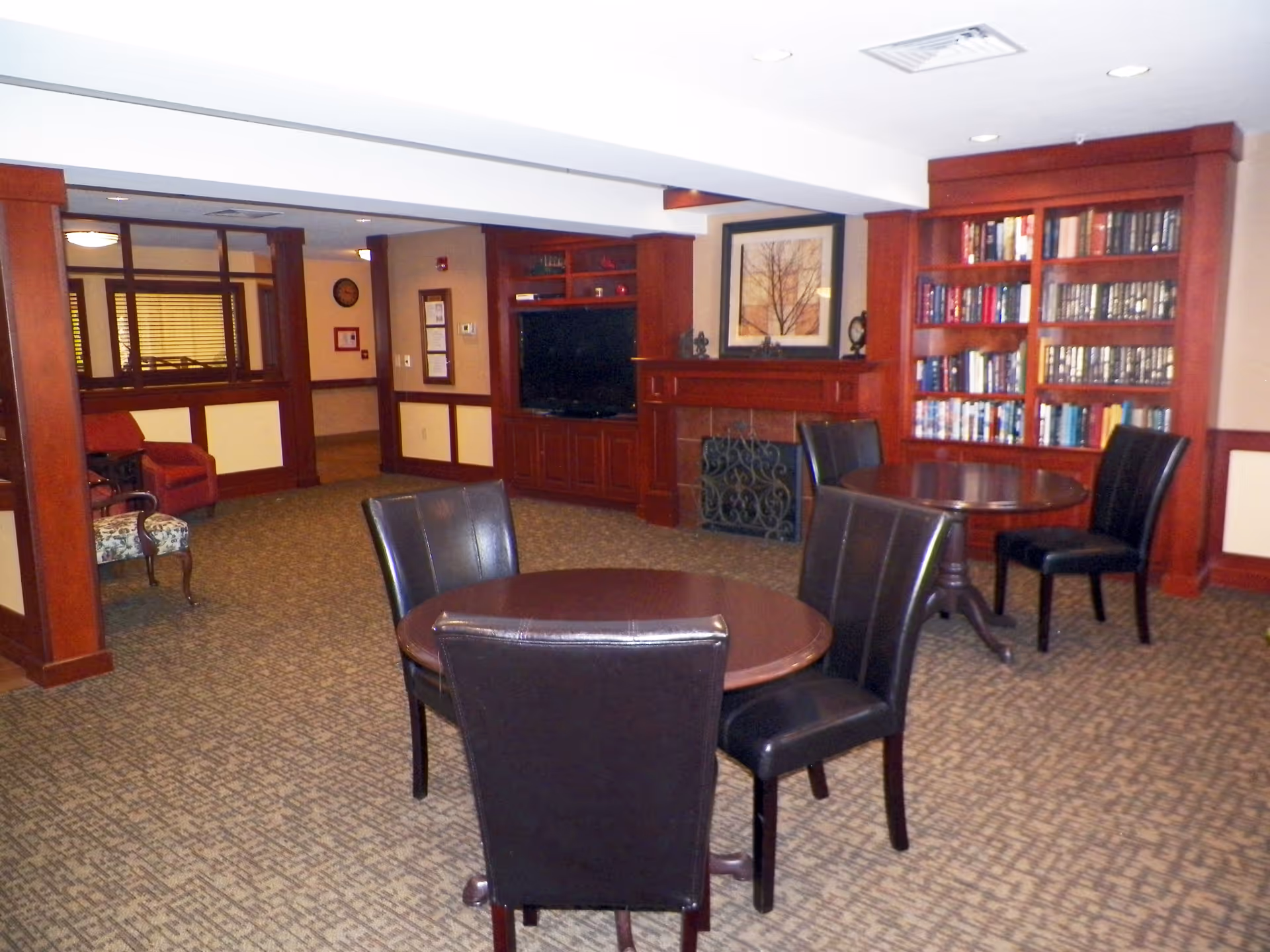 A cozy common area with carpeted floor featuring two round wooden tables each surrounded by black leather chairs. There is a built-in wooden bookshelf filled with books, a fireplace with a decorative screen, and a flat-screen TV mounted in a wooden entertainment center. The room has warm wood paneling and trim, with additional seating including an upholstered armchair and a patterned chair near the back.