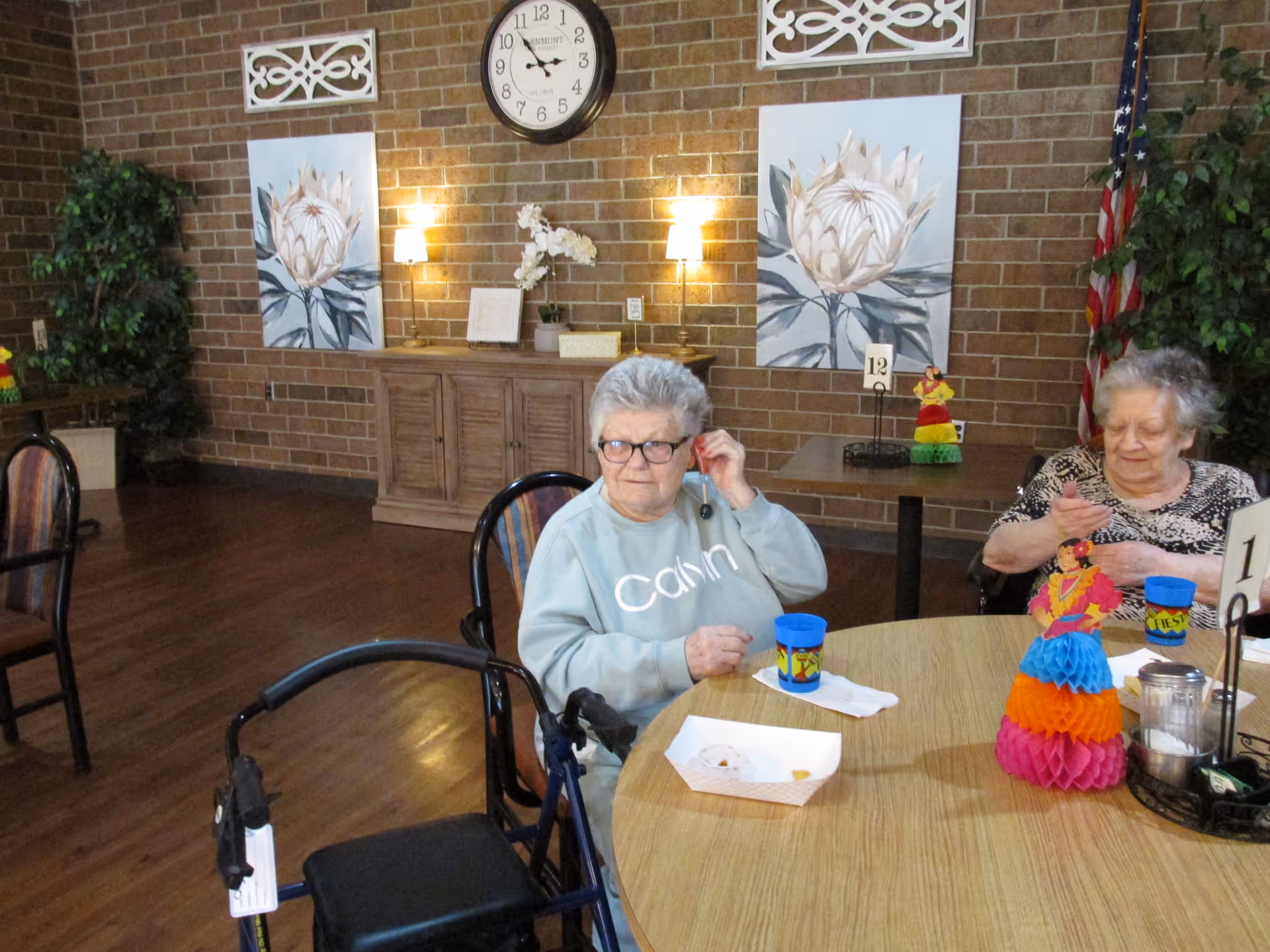 Two elderly women sitting at a round wooden table in a common area with brick walls. One woman is wearing glasses and a light blue sweatshirt with the word 'Calm' on it, and the other woman is wearing a patterned top. The table has colorful decorations, cups, and a small food tray. Behind them are two large floral paintings, a clock, wall sconces, and some plants, including an American flag in the corner.