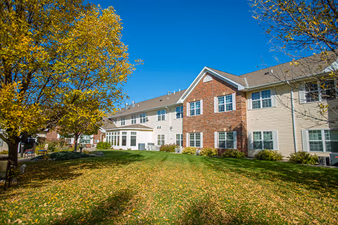 Two-story brick-and-siding assisted living building with a grassy lawn and autumn trees under a clear blue sky.