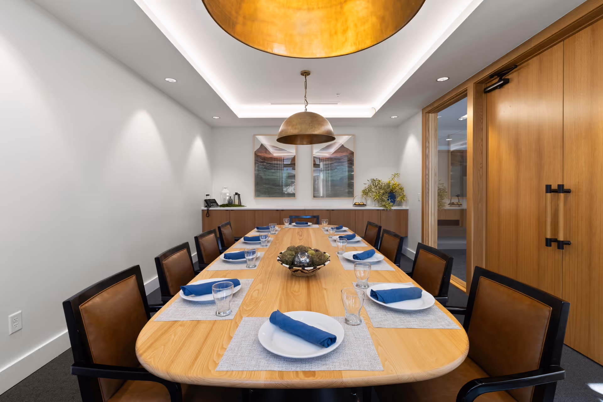 A modern dining room with a long wooden table set for eight people. Each place setting includes a white plate with a blue cloth napkin and a clear drinking glass on a light gray placemat. The room features brown leather chairs, a large round metallic pendant light hanging from the ceiling, and two framed artworks on the far wall above a wooden sideboard with decorative items and a telephone.