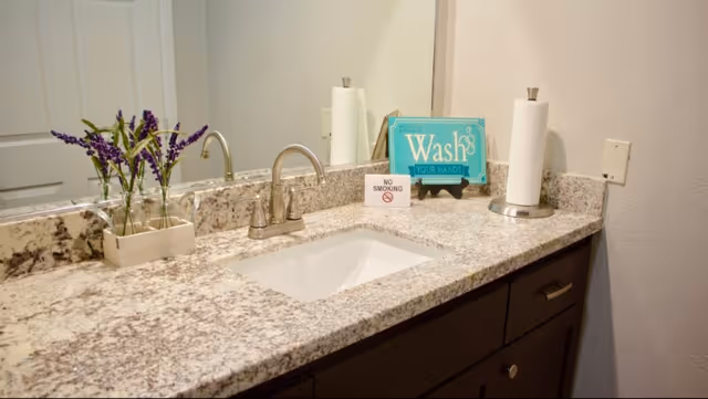 A bathroom countertop with a sink, silver faucet, and a large mirror. On the countertop, there is a small vase with purple flowers, a paper towel holder with a roll of paper towels, a small sign that says 'NO SMOKING', and a decorative sign that says 'Please Wash Your Hands'.
