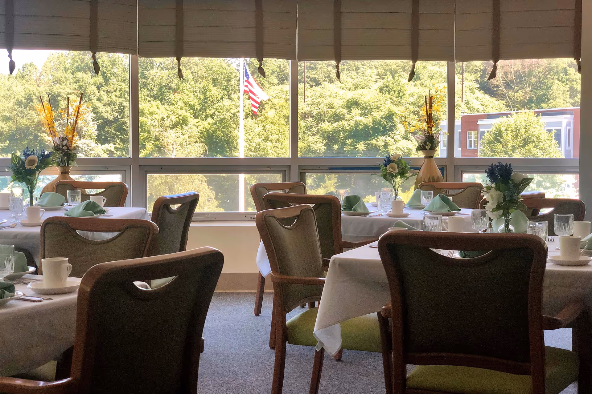 Sunlit dining room with tables set with white tablecloths, green napkins, floral centerpieces and chairs in front of large windows showing trees and an American flag.