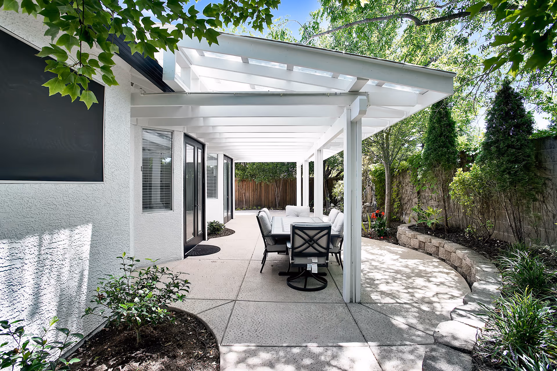 Outdoor patio area at Foothills Senior Care with a white pergola covering a dining table and cushioned chairs. The patio is surrounded by greenery including trees, shrubs, and a stone retaining wall.