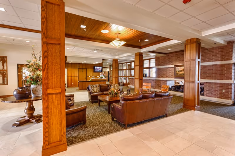 A spacious senior living facility common area with leather sofas and armchairs arranged around a wooden coffee table on a patterned carpet. The room features wooden pillars, a wooden ceiling accent with a chandelier, brick walls, and a reception desk in the background. There are decorative elements including a large floral arrangement on a round wooden table and framed artwork on the walls.