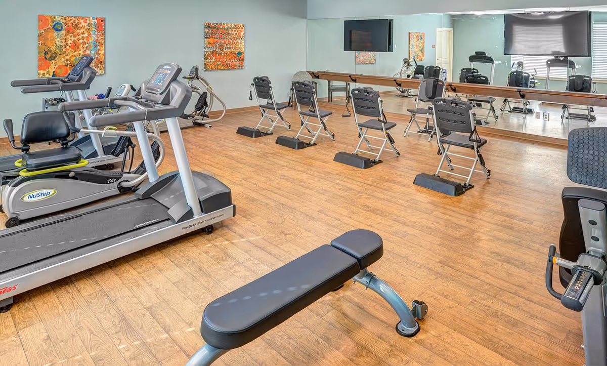 A fitness room with exercise equipment including treadmills, stationary bikes, and a workout bench. The room has wooden flooring, a large mirror on one wall, colorful artwork, and a wall-mounted television.