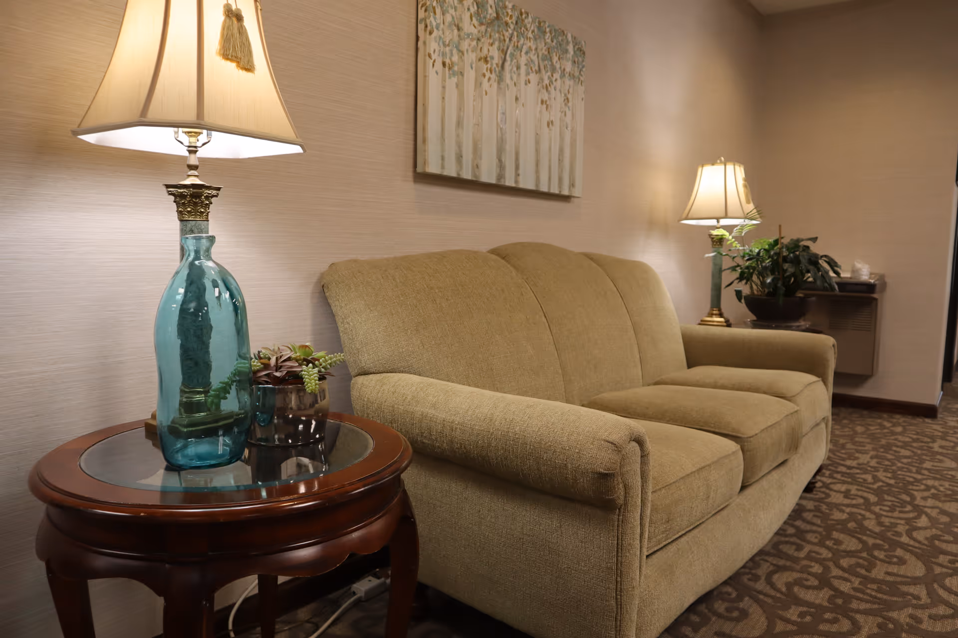A cozy living room area featuring a beige upholstered sofa, a wooden side table with a glass top holding a decorative blue glass lamp and a small plant. There is a second lamp and a potted plant on a small wall-mounted shelf in the background. A painting with a nature theme hangs on the wall above the sofa.