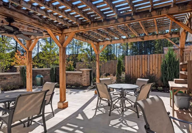 Outdoor patio area with a wooden pergola overhead, featuring several metal and mesh chairs around round tables on a concrete floor. The space is surrounded by a wooden fence and landscaped with small trees and shrubs under a clear blue sky.
