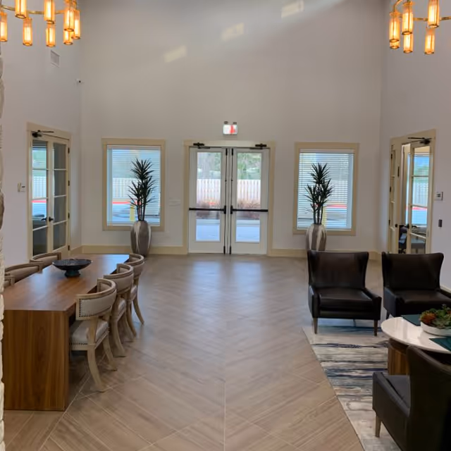 Bright lobby-style common room with a long dining table and chairs on the left, lounge seating on the right, potted plants, and double glass entrance doors centered at the back.