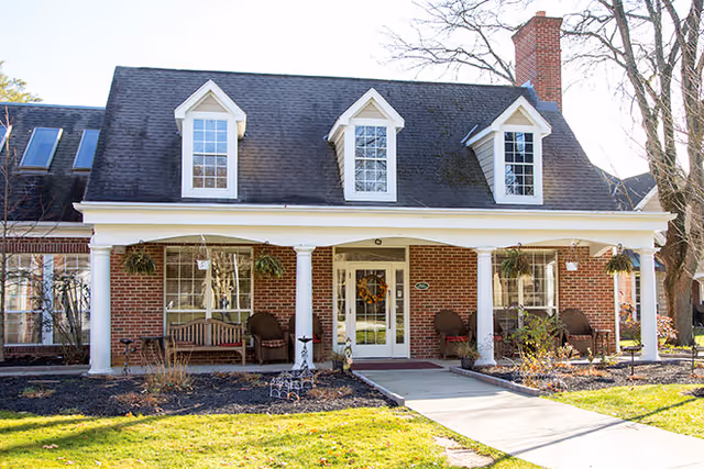 Front exterior view of a brick building with a covered porch supported by white columns. The porch has seating including chairs and a bench, hanging plants, and a wreath on the glass front door. The building has three dormer windows on the roof and a chimney on the right side.