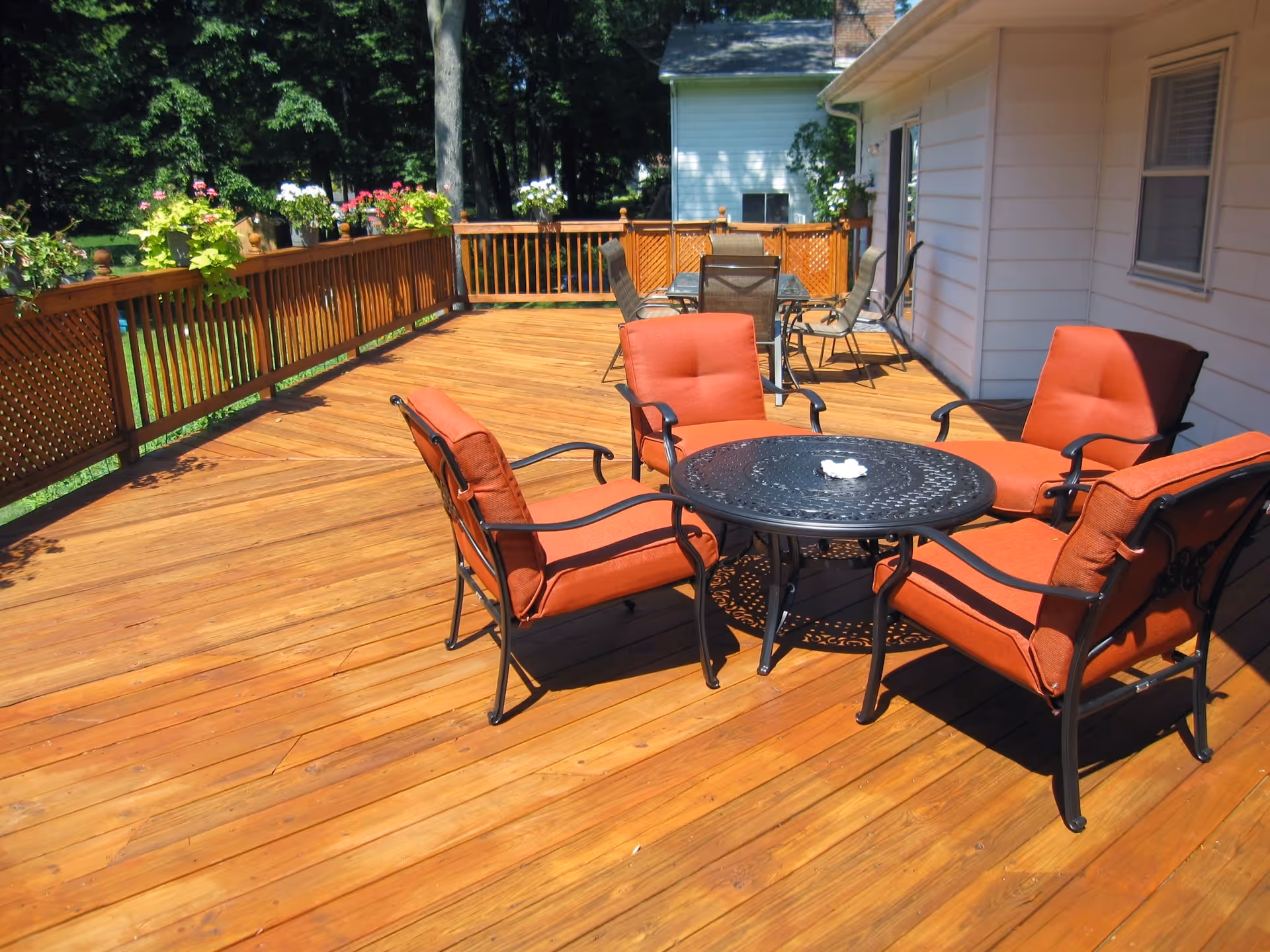 Outdoor wooden deck with a round black metal table surrounded by four orange cushioned chairs. In the background, there is another table with several chairs and flower pots hanging on the wooden railing. Trees and greenery are visible beyond the deck.