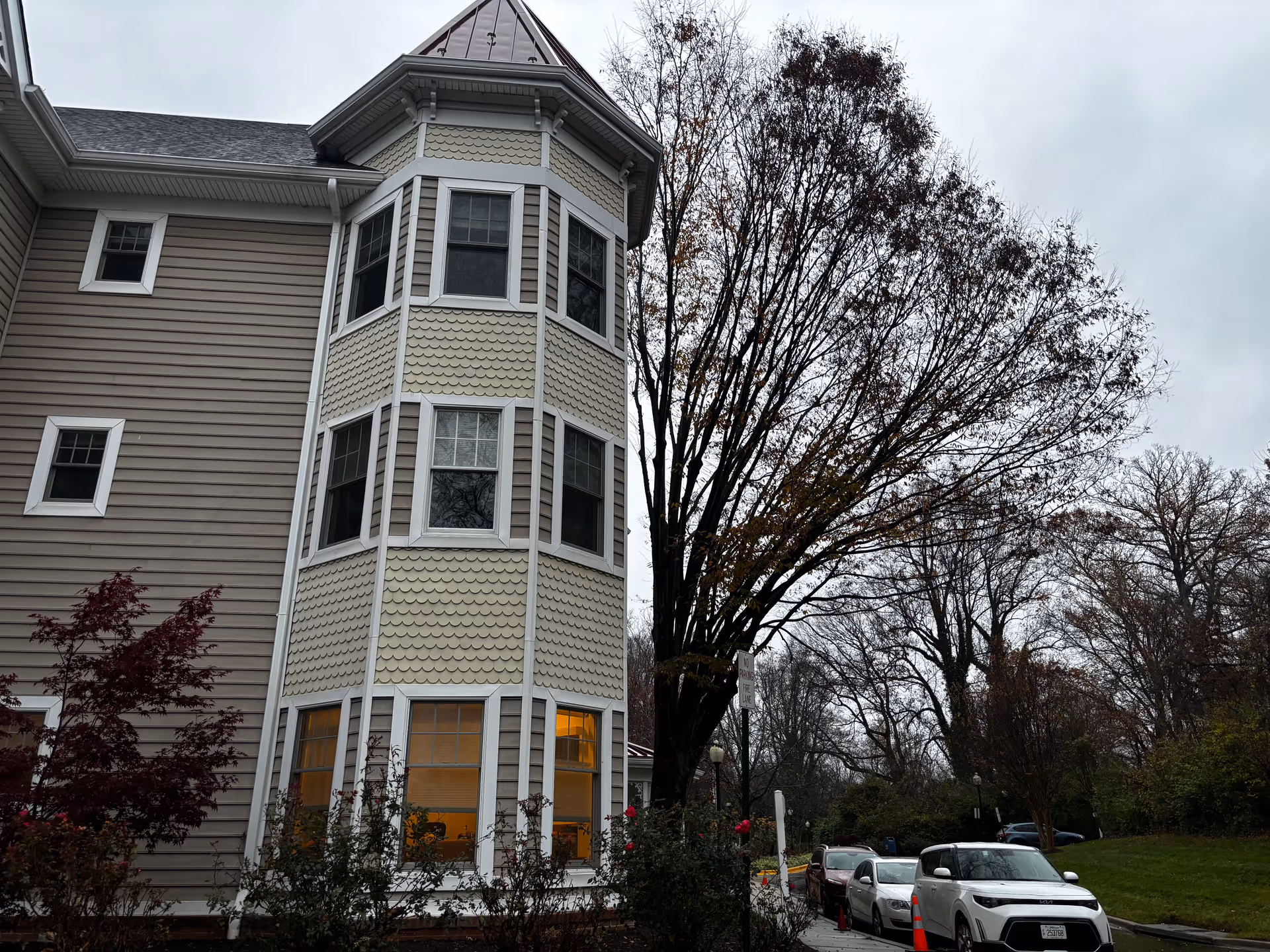 Exterior view of a multistory senior living building with bay windows, a large leafless tree, and cars parked along the driveway on a cloudy day.