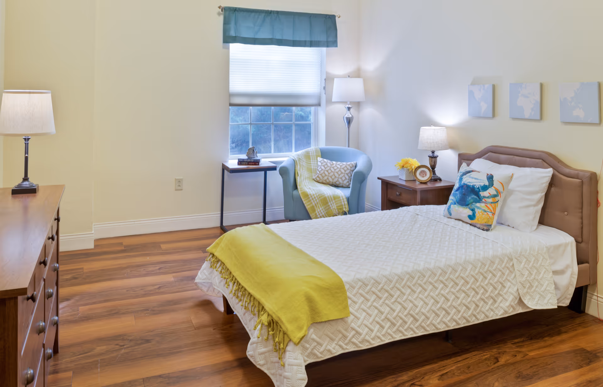 A cozy bedroom with a single bed covered in a white textured bedspread and a yellow throw blanket. The bed has decorative pillows, including one with a colorful crab design. Next to the bed is a wooden nightstand with a lamp, a small clock, and a vase of yellow flowers. A comfortable blue armchair with a yellow and white throw blanket and a patterned pillow sits near a window with a blue valance and white blinds. A wooden dresser with a lamp is on the left side of the room. The floor is wooden, and the walls are painted light yellow with three small world map artworks hanging above the bed.