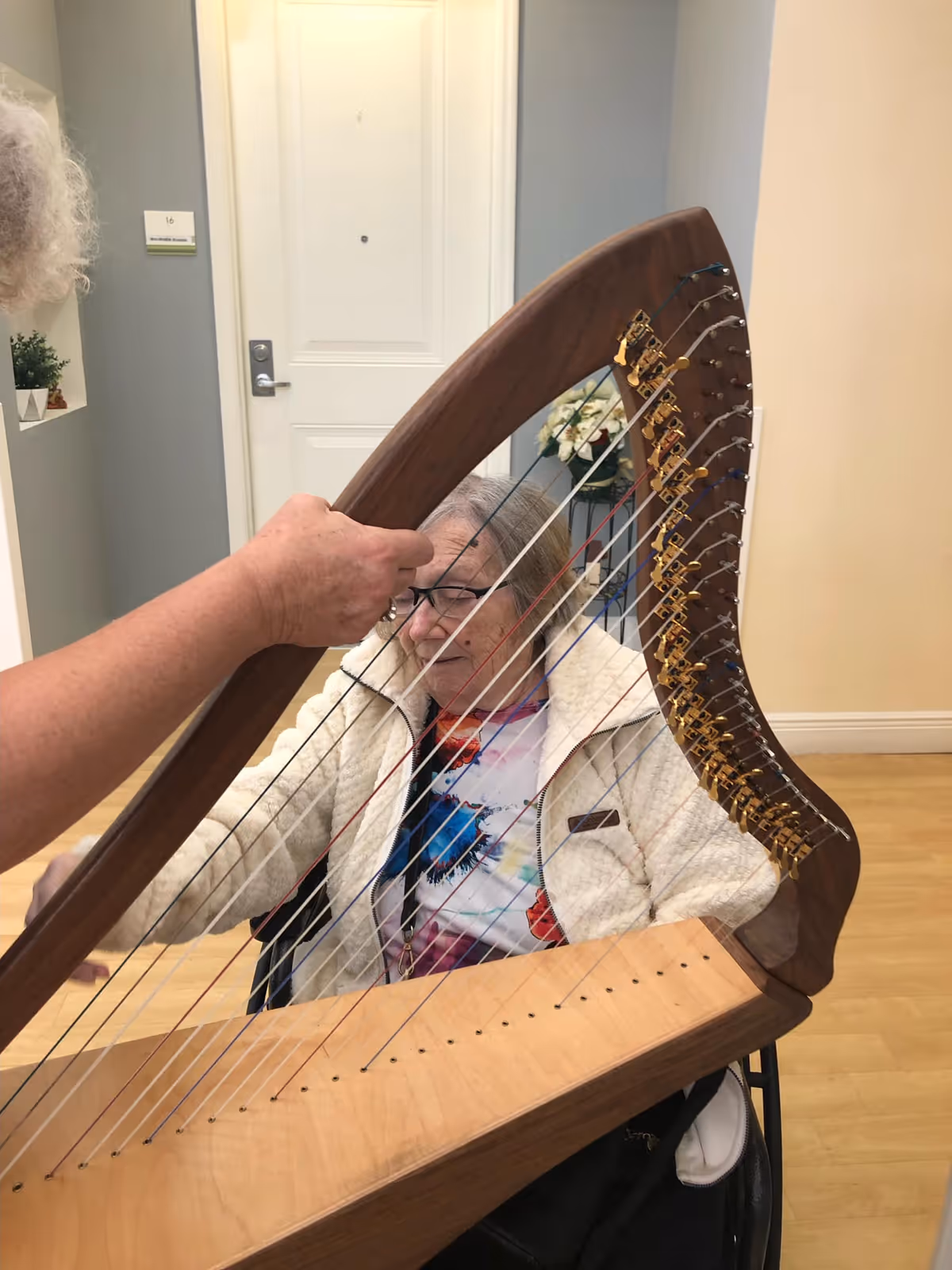 An elderly woman seated and playing a wooden harp with the assistance of another person's hand in a well-lit room with a white door and light-colored walls in the background.