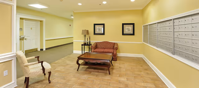 A hallway area in a senior living facility with a beige tiled floor and yellow walls. On the right side, there is a large bank of mailboxes. In the center, there is a wooden coffee table in front of a red patterned loveseat, with a side table holding a lamp and decorative items. On the left, there is a single upholstered armchair. The hallway extends into the background with doors and a carpeted floor.