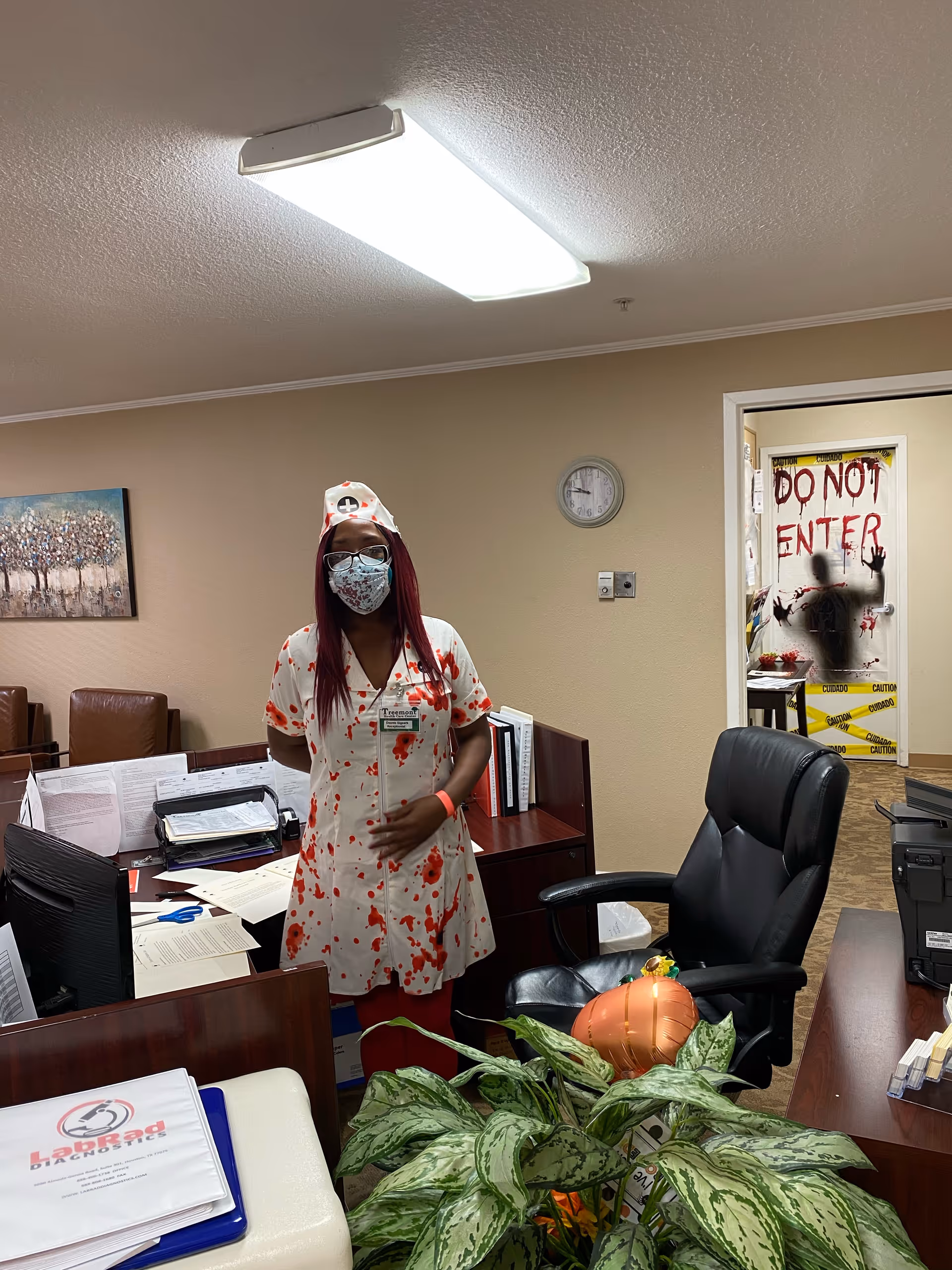 A person dressed in a nurse costume with fake blood stains and a face mask stands behind a desk in an office setting. The desk has papers, folders, and office supplies on it. Behind the person, a door is decorated with a spooky theme, including the words 'DO NOT ENTER' written in red and a silhouette of a person pressing against the door, with caution tape around the door frame.