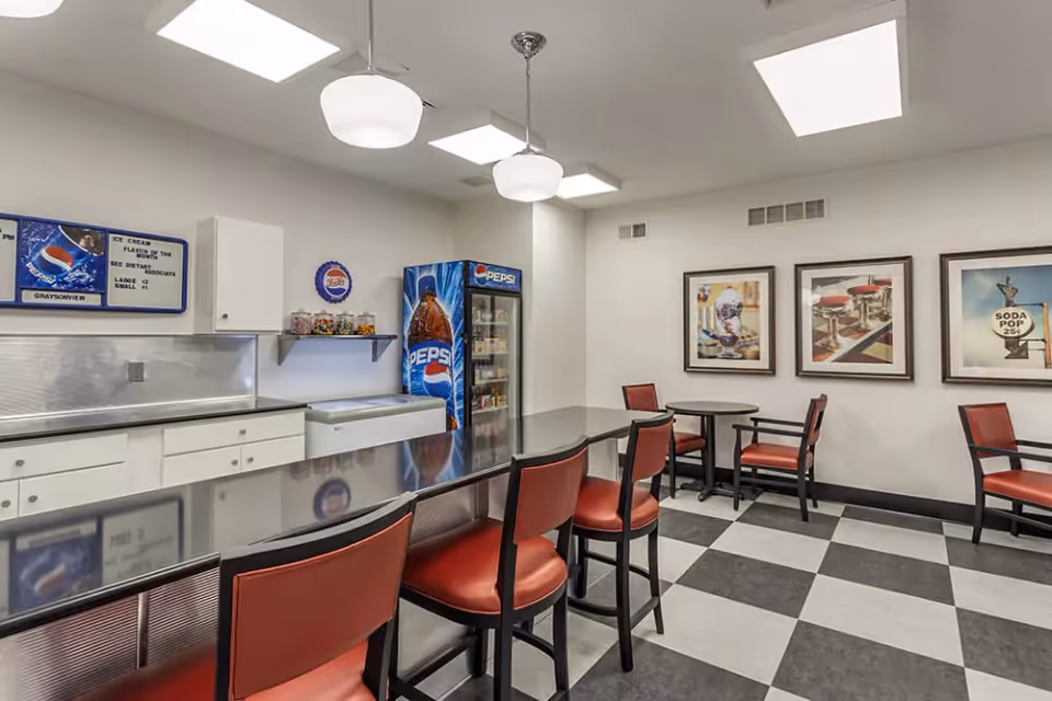 Interior view of a dining area with a long counter and red cushioned chairs. The room features a checkered black and white floor, white walls with three framed pictures, a Pepsi vending machine, and a small freezer. Overhead lighting includes two pendant lights and ceiling panels.