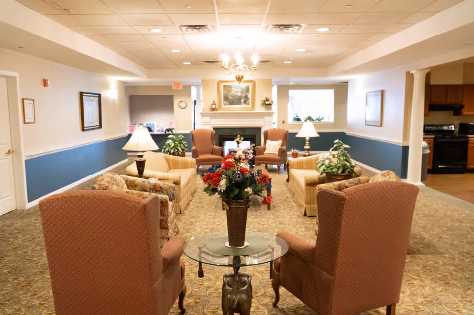 A cozy senior living facility common area with multiple upholstered armchairs and sofas arranged around a glass coffee table with a flower arrangement. The room features a fireplace with a painting above it, table lamps, and plants. The walls are painted beige with a blue accent panel, and the carpet has a floral pattern. In the background, there is a kitchen area visible to the right.