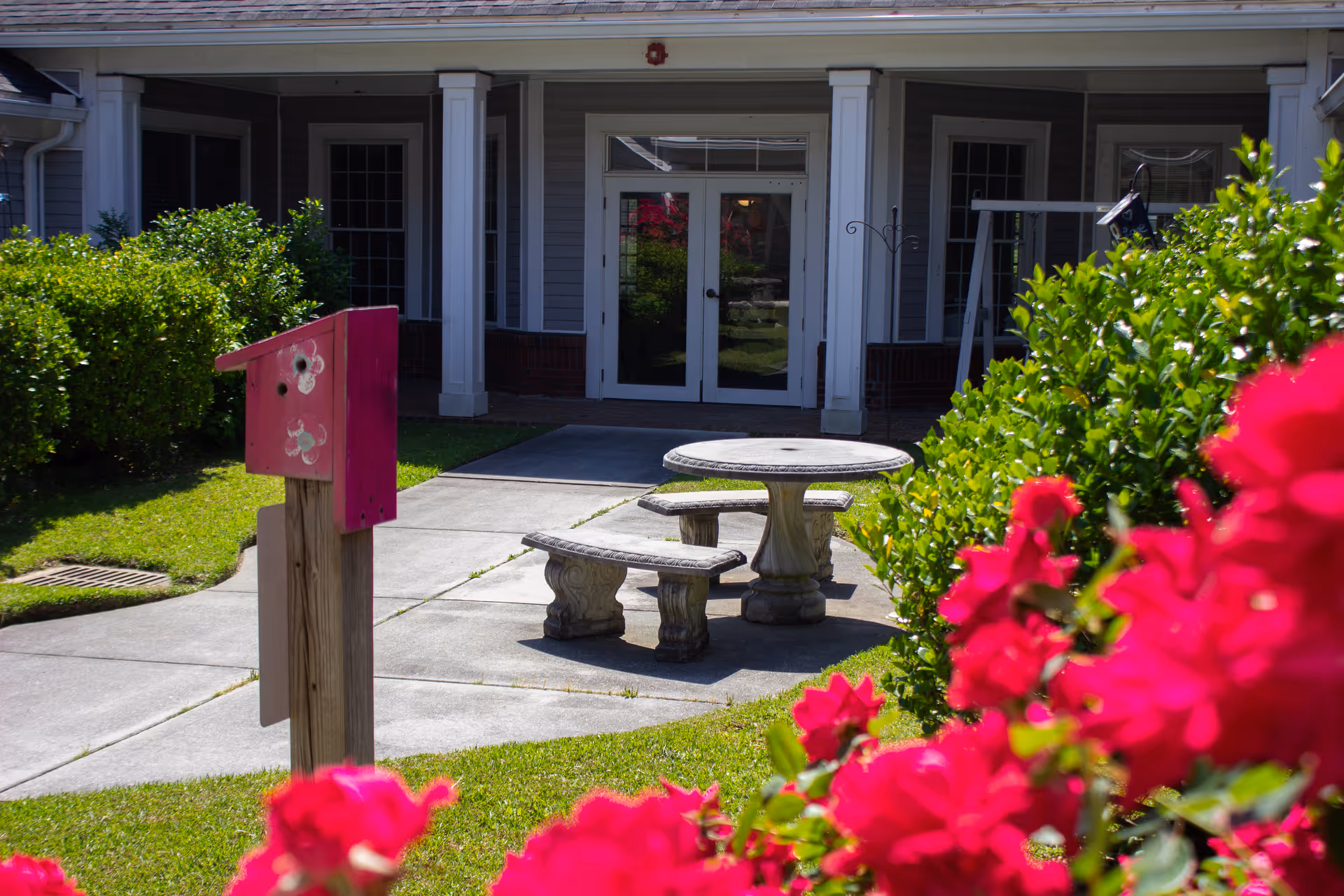 Outdoor area of a senior living facility with a concrete round table and two curved benches on a paved patio. There are green bushes and vibrant red flowers in the foreground, and a building entrance with double glass doors and white columns in the background.