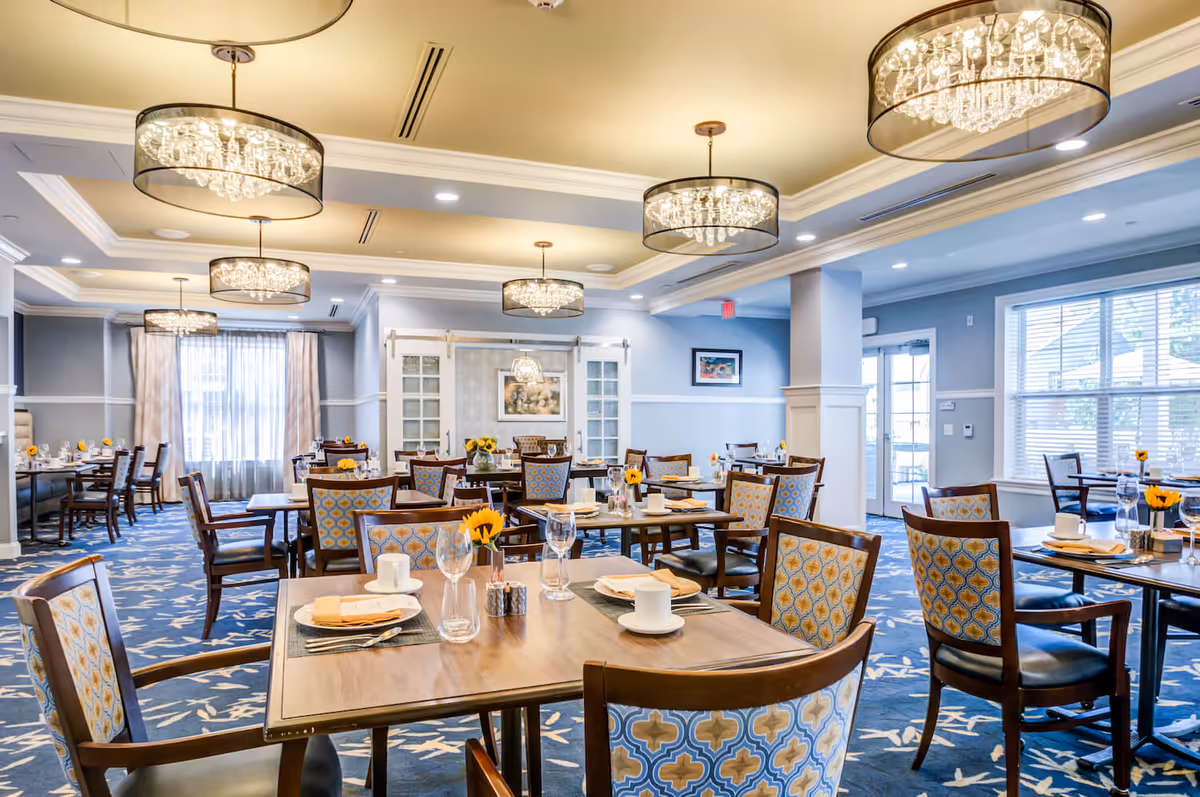 A bright and elegant dining room with multiple wooden tables and chairs featuring patterned upholstery. Each table is set with plates, cups, glasses, and napkins, and decorated with small vases holding yellow flowers. The room has large windows with blinds and curtains, blue carpet with a white floral pattern, and several modern chandeliers hanging from the ceiling. Artwork is displayed on the walls, and there is a door leading outside.