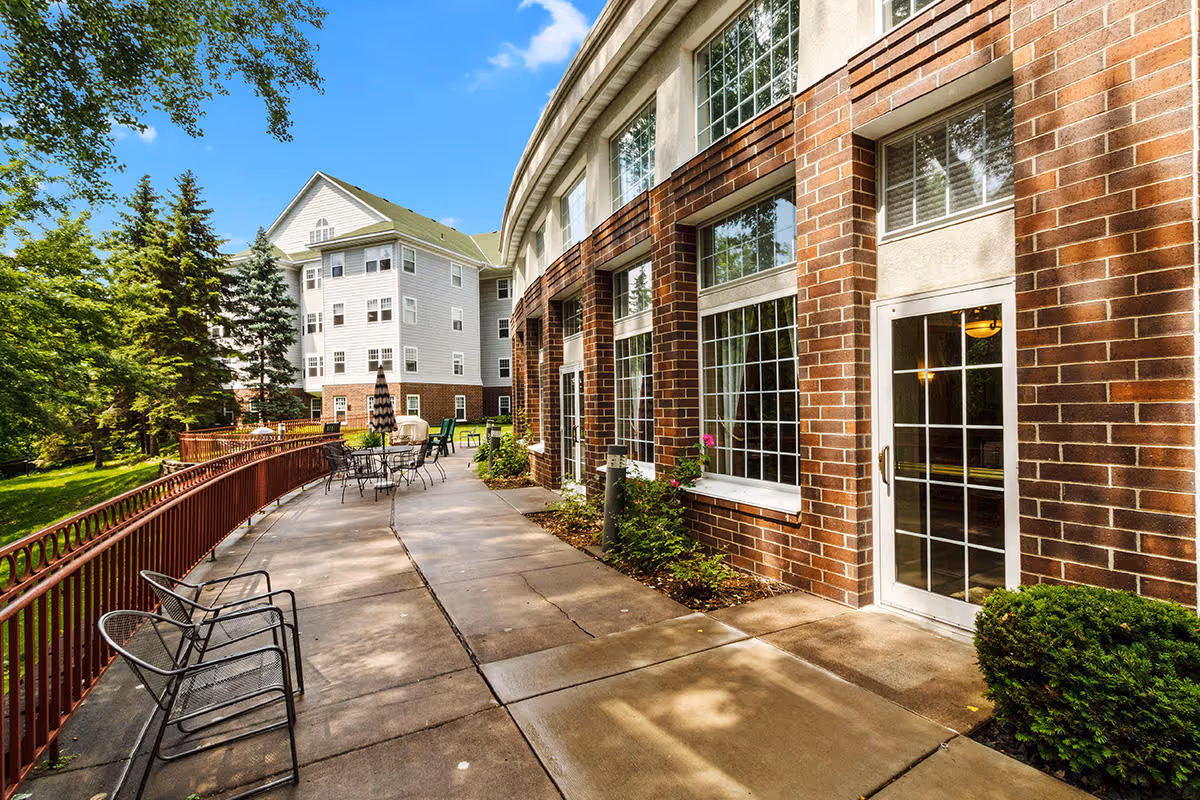 Outdoor patio area of a senior housing facility with metal chairs and tables, a red railing, and a brick building with large windows and a glass door. Trees and greenery surround the area under a clear blue sky.