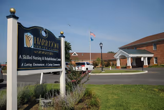 Exterior view of Harrison Senior Living of Gloucester, showing the entrance with a covered drop-off area, a flagpole with an American flag, and a sign in the foreground indicating it is a skilled nursing and rehabilitation center.