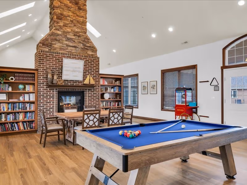 A spacious recreational room with a blue felt pool table in the foreground, a brick and stone fireplace with a wooden mantel in the background, flanked by bookshelves filled with books and decorative items. There is a wooden table with chairs near the fireplace, a popcorn machine against the wall, and windows with blinds letting in natural light.