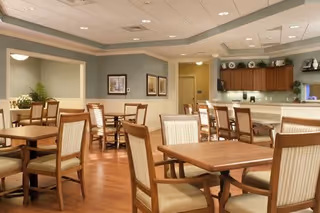 A spacious dining room with multiple wooden tables and chairs arranged neatly on a polished wooden floor. The walls are painted light green and decorated with framed pictures. There is a counter area with cabinets and a few decorative plates on display. The ceiling has recessed lighting.