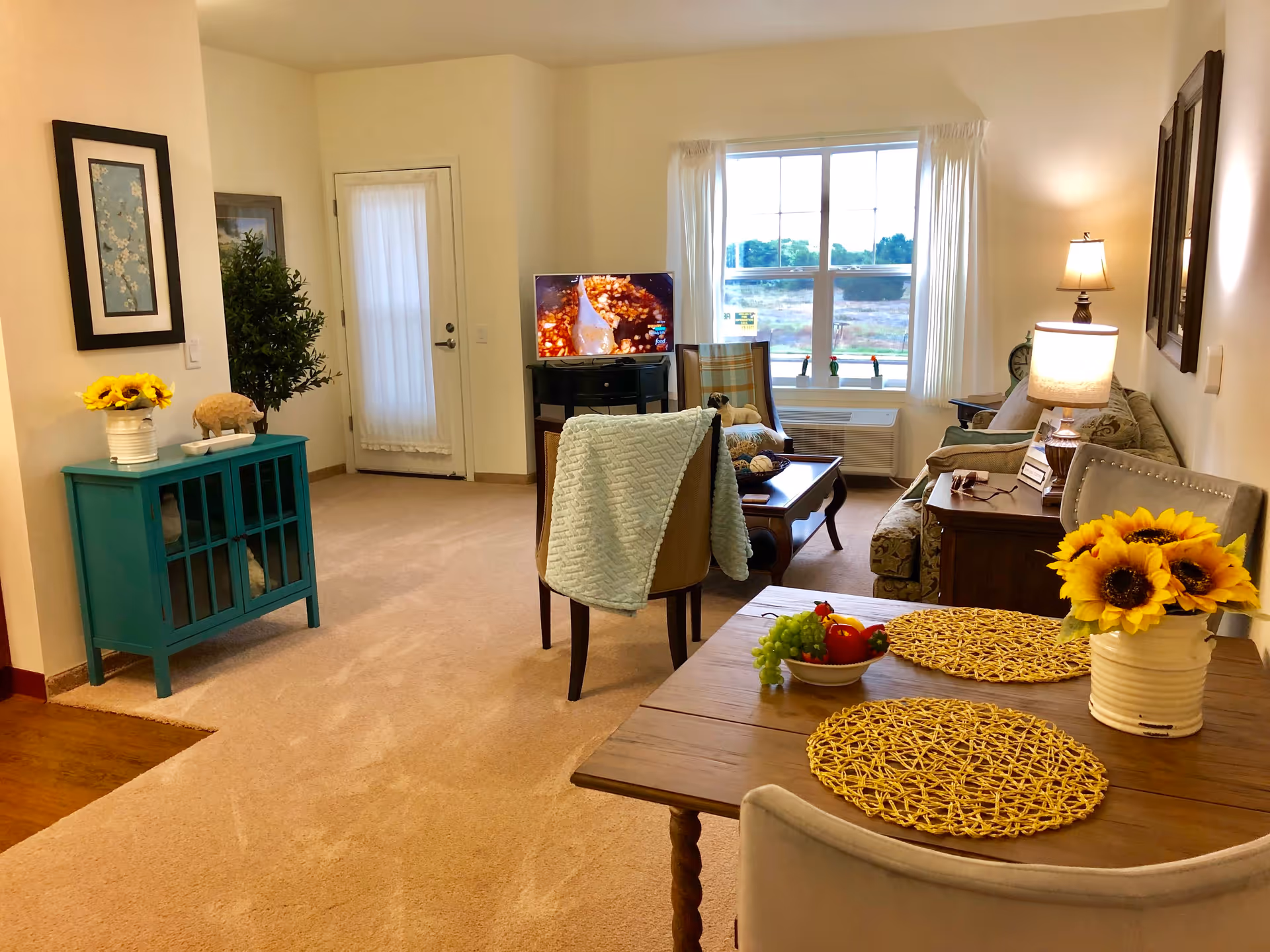 Sunlit living room and dining area with a sofa, TV, coffee table, and a dining table decorated with sunflowers.