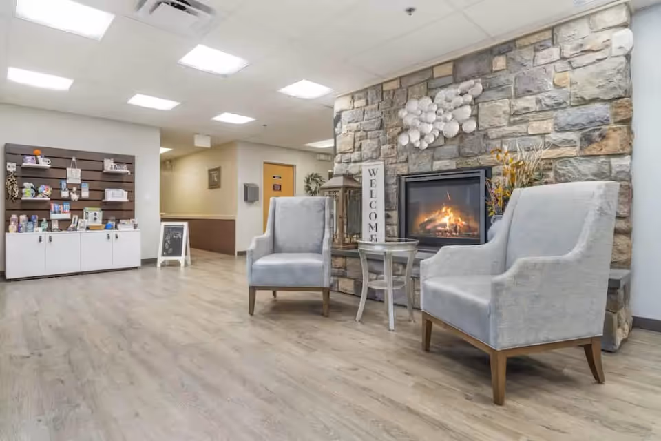 A cozy seating area in a senior living facility with two gray armchairs facing a stone fireplace with a fire burning. A small round table is placed between the chairs, and a vertical sign that says 'WELCOME' is next to the fireplace. The room has light wood flooring, bright ceiling lights, and a display shelf with various items on the left side.
