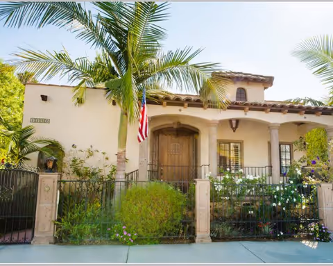 Front exterior of a Spanish-style house with a wooden arched door, columns, palm trees, an American flag, and a low iron fence.