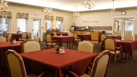 Dining room with multiple tables covered in burgundy tablecloths, upholstered chairs, chandeliers, and a buffet/service area at the back.