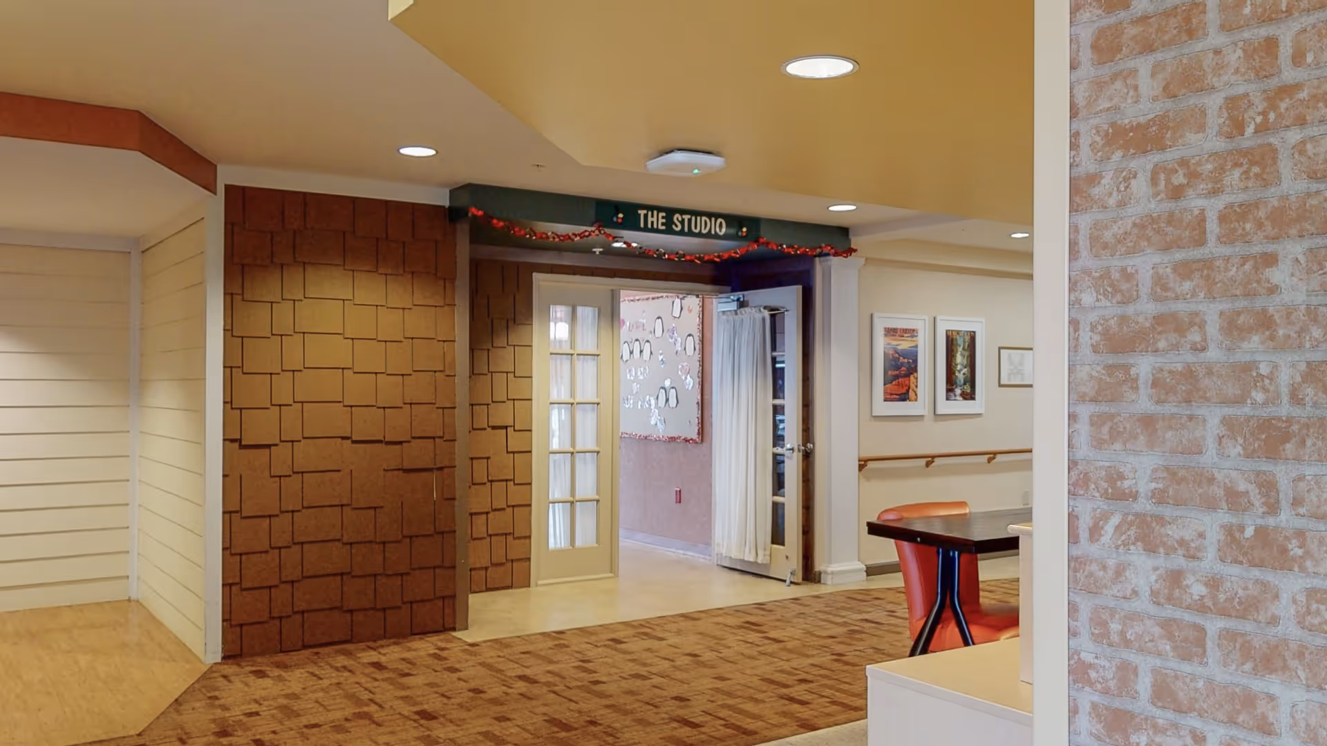 Interior view of a senior living facility hallway leading to a room labeled 'The Studio'. The hallway features a mix of wall textures including white paneling, brown shingle-like tiles, and exposed brick. There is a table with red chairs partially visible on the right side, and framed pictures on the wall in the background.