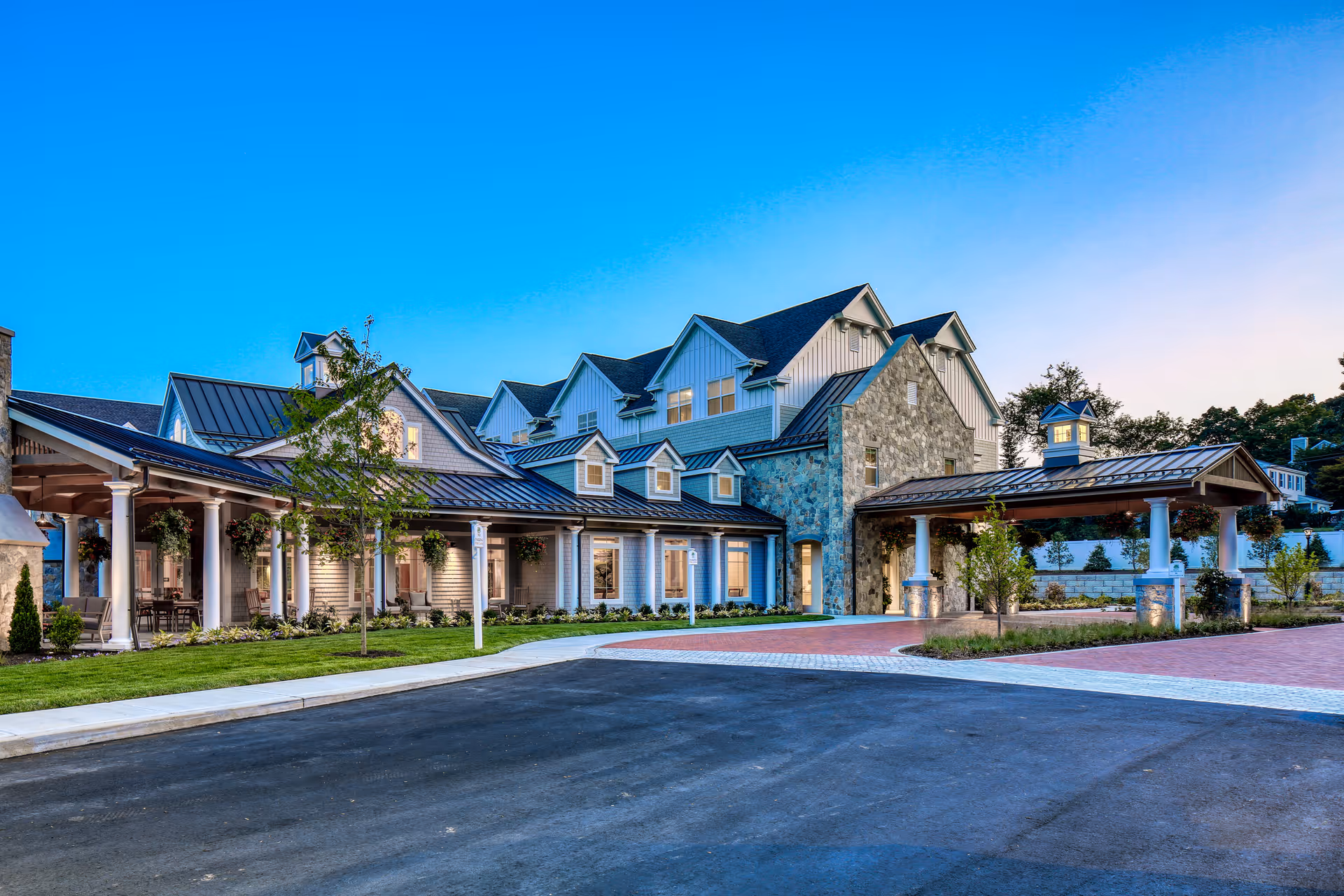 Exterior view of a senior living facility building at dusk with a covered entrance, stone and siding exterior, multiple windows, and landscaped surroundings.