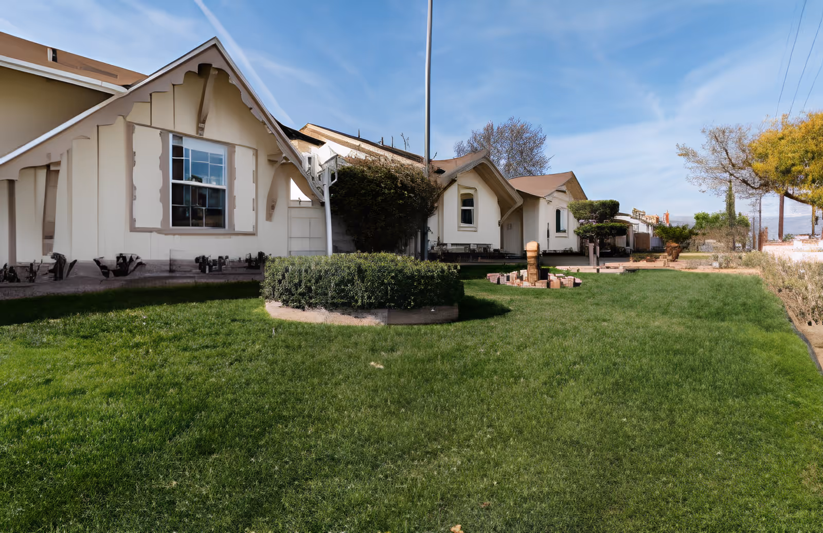 Single-story cream-colored building front with a green lawn, shrubs, a flagpole, and blue sky.