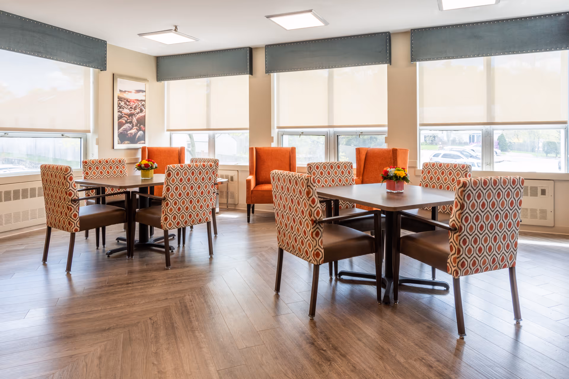 A bright dining area with two square tables, each surrounded by four patterned chairs with a red and beige geometric design. There are also two orange armchairs near the windows. The room has large windows with beige roller shades, wooden flooring, and a framed picture on the wall.