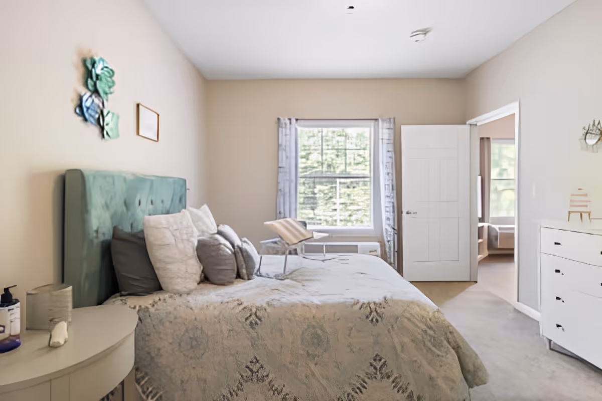 A cozy bedroom with a large bed featuring a green upholstered headboard and multiple pillows. There is a small round bedside table with a lamp and hand sanitizer. A window with patterned curtains lets in natural light, and a white dresser is positioned against the opposite wall. The room has light-colored walls and carpeted flooring, with an open door leading to another room.