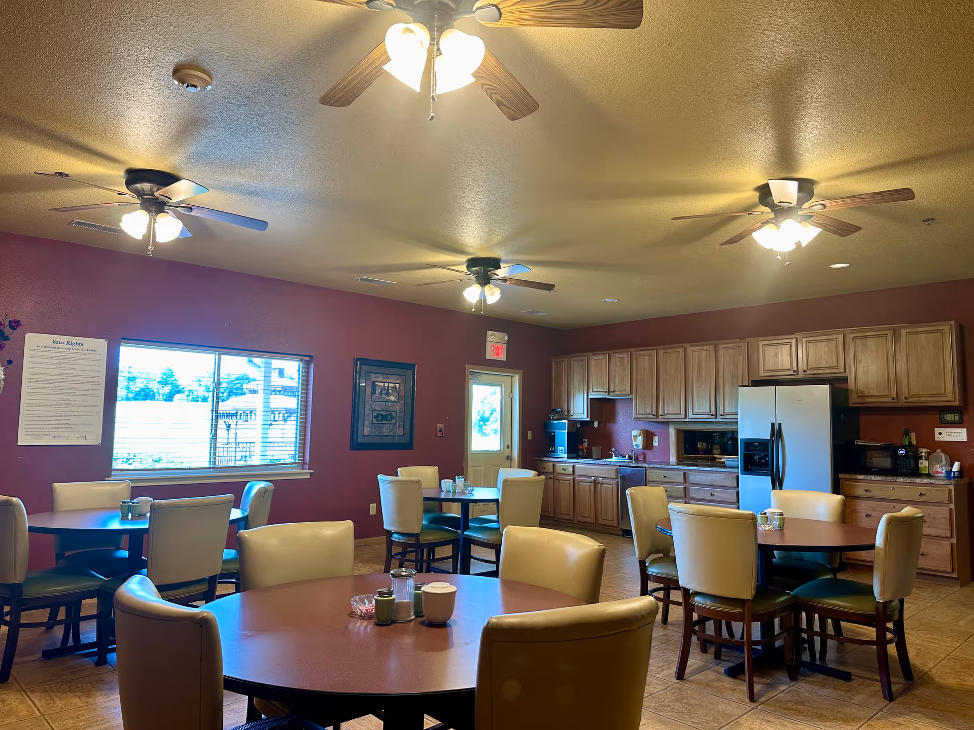 Communal dining area with round tables and chairs and a kitchenette with cabinets and a refrigerator along the back wall.