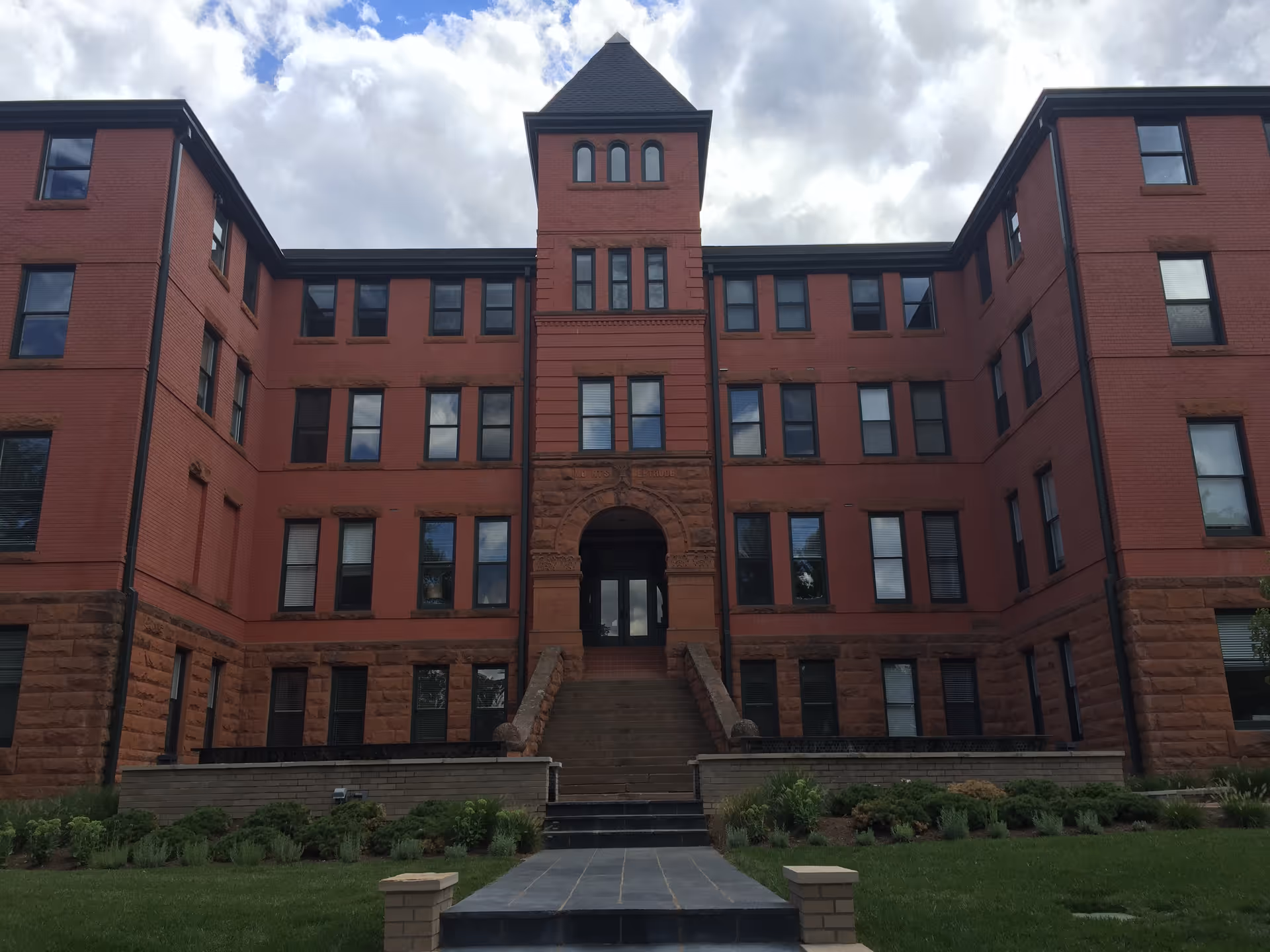 Front exterior view of a large, multi-story brick building with a central tower and arched entrance, surrounded by a well-maintained lawn and shrubbery under a partly cloudy sky.