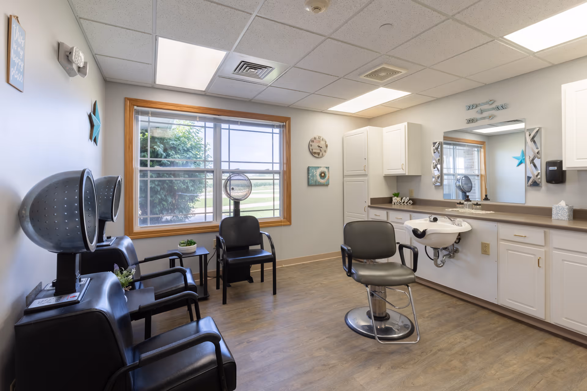 Small salon in a senior facility with a styling chair and sink, hooded hair dryers, and waiting chairs by a window.