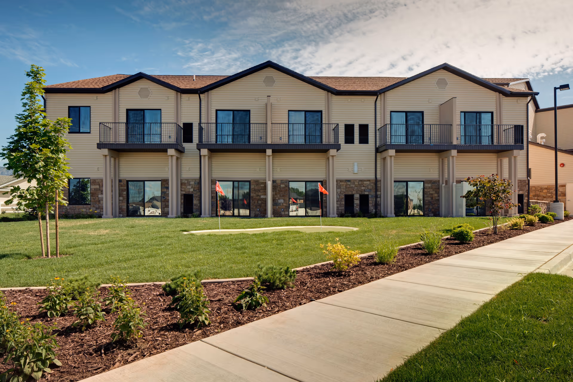 Exterior view of a two-story building with beige siding and stone accents, featuring balconies on the upper floor and large windows on the ground floor. In front of the building is a well-maintained lawn with a small putting green marked by red flags, bordered by a sidewalk and landscaped with small plants and shrubs.
